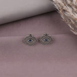 A pair of silver-toned evil eye stud earrings with blue stone centers, displayed on a dusty mauve surface with dried pampas grass in the corner.