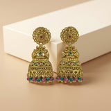 A pair of ornate gold-toned Indian jhumka earrings with colorful green, pink, and blue stone accents, displayed on a beige surface with a white geometric block in the background.