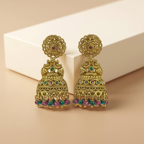 A pair of ornate gold-toned Indian jhumka earrings with colorful green, pink, and blue stone accents, displayed on a beige surface with a white geometric block in the background.