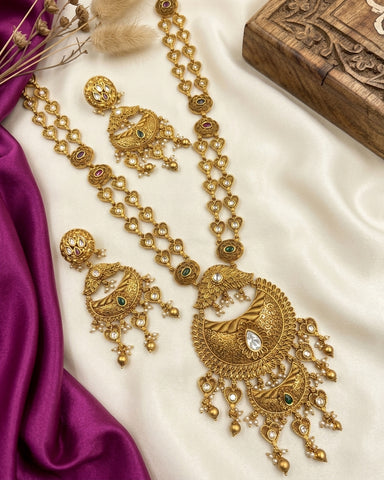 An ornate gold-toned traditional necklace and matching earrings laid out on a cream satin fabric, with a purple satin ribbon and a carved wooden box as styling props.