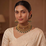 A woman wearing an ornate gold-toned traditional necklace with green bead accents and matching dangling earrings, paired with a light cream saree in a softly lit indoor setting.