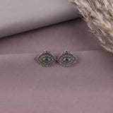 A pair of silver-toned geometric stud earrings with green oval centers, displayed on a dusty mauve surface with dried grass in the corner.