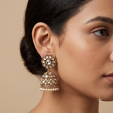 A close-up of a woman's ear wearing an ornate gold-toned jhumka earring with white stones and pearl-like beads, against a warm beige background.