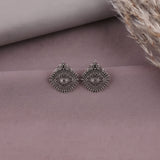 A pair of silver-toned geometric stud earrings with intricate filigree detailing and a central eye motif, displayed on a textured mauve surface with neutral props.
