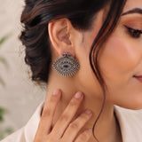 A close-up of a woman wearing a large silver-toned traditional Indian jhumka earring with a blue stone and silver bead detailing, with her hand resting on her neck.