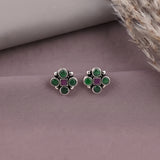 A pair of silver stud earrings with five green round stones and a central square purple stone, displayed on a textured mauve surface with dried grass in the corner.