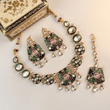 Gold-plated traditional Indian jewelry set featuring a necklace, matching earrings, and a dangling pendant, decorated with green and pink enamel, clear stones, and teardrop crystals. A small ornate cream-colored jewelry box with gold floral detailing is partially visible in the top left corner on a light beige background.