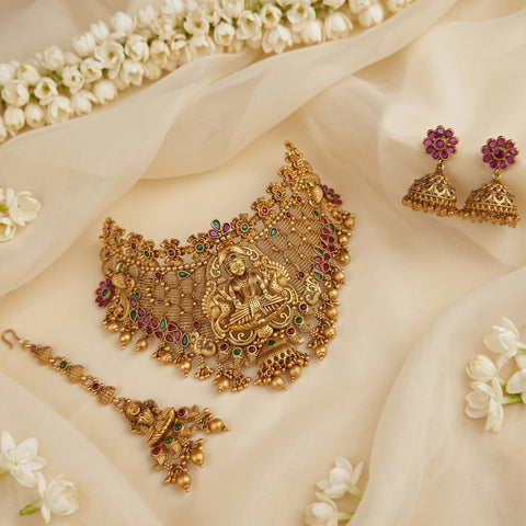 An ornate gold-toned traditional jewelry set arranged on cream fabric with white flowers. The set includes a large necklace with a central deity motif and colorful stone accents, matching earrings, and a dangling bracelet or maang tikka.