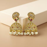 A pair of gold-plated Indian jhumka earrings adorned with clear pavé stones, small red gemstones, green accent stones, and dangling white faux pearls, displayed on a beige background.