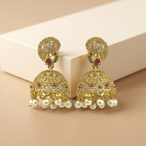 A pair of gold-plated Indian jhumka earrings adorned with clear pavé stones, small red gemstones, green accent stones, and dangling white faux pearls, displayed on a beige background.