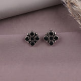 A pair of silver-toned floral stud earrings with black round and square stones, displayed on a textured mauve surface with a neutral prop in the background.