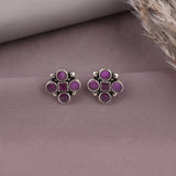 A pair of silver-toned stud earrings with five purple gemstones arranged in a flower-like cluster, displayed on a dusty mauve surface with dried grass in the corner.