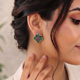A close-up of a woman wearing a silver-toned four-petal flower earring with green and magenta stones, shown on her ear and neck.