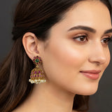 A close-up of a woman's ear wearing a gold-toned traditional Indian jhumka earring embellished with pink stones, green stones, and small white pearl-like beads.