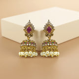A pair of ornate gold-toned jhumka earrings with oval purple gemstones, clear rhinestones, and white pearl drops, displayed on a beige surface in front of a cream-colored box.