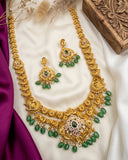 Gold-toned traditional Indian jewelry set featuring a necklace, earrings, and a matching bracelet, decorated with green stone accents and small white pearl-like beads, arranged on a white satin fabric with purple silk and a carved wooden box.