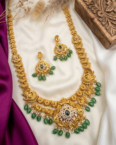 Gold-toned traditional Indian jewelry set featuring a necklace, earrings, and a matching bracelet, decorated with green stone accents and small white pearl-like beads, arranged on a white satin fabric with purple silk and a carved wooden box.