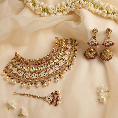 An ornate Indian jewelry set arranged on draped cream fabric with white flowers. The set includes a gold-toned choker necklace with white enamel inlays, red and green stone accents, dangling pearl-like beads, matching dangle earrings, a maang tikka, and a small chain connector.
