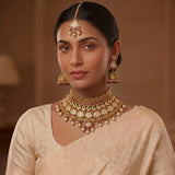 A woman wearing traditional Indian jewelry, including a gold-toned maang tikka, matching earrings, and a statement choker necklace with gold coins, pearls, and pink stones, paired with a light beige saree.
