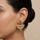 A close-up of a woman's ear wearing a gold-toned traditional Indian chandelier earring decorated with red and green stones, white faux pearls, and a dangling light green bead.