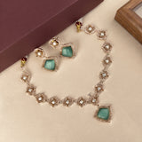 A gold-tone jewelry set featuring a necklace and matching earrings with square mint green cabochon stones surrounded by clear crystals, displayed on a beige surface with a dark brown box in the background.