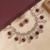 Gold-plated statement jewelry set featuring a necklace, matching earrings, and a bracelet, embellished with deep red teardrop gemstones and clear round crystals on a beige velvet surface.