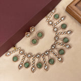 Gold-plated Indian ethnic jewelry set featuring a necklace and matching earrings with oval green stones and clear teardrop crystals, displayed on a beige surface next to a maroon box.