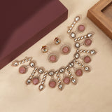 A jewelry set featuring a statement necklace, matching earrings, and stud earrings in rose gold with pink gemstone accents and clear rhinestones, arranged on a beige surface next to a dark maroon box.