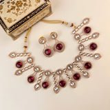 A traditional Indian jewelry set featuring a statement necklace, matching earrings, and a decorative gift box. The jewelry is made of rose gold-toned metal, adorned with small white pearls and deep red gemstones, displayed on a light beige background.