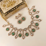 An ornate gold and silver jewelry set featuring a statement necklace and matching earrings with green stone accents, displayed next to a decorative cream and gold clutch bag on a light beige background.