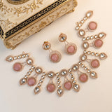 Gold-toned traditional Indian jewelry set featuring a necklace, earrings, and a matching decorative box with pink stone accents and clear crystal details, arranged on a light cream background.