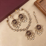 Gold-toned Indian jewelry set featuring a necklace, earrings, and a bracelet with red and green enamel details and clear stones, arranged on a beige surface with a maroon box corner in the background.