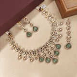 Gold-plated Kalka jewelry set with green stone accents, including a necklace, earrings, and a bracelet, arranged on a beige surface with a brown box in the background.