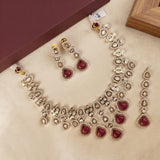 Gold-plated Indian Kundan jewelry set featuring a necklace, matching earrings, and a dangling choker, decorated with clear crystals and deep red heart-shaped stones, displayed on a beige surface with a brown box in the background.