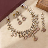 A traditional Indian jewelry set featuring a necklace, earrings, and a dangling maang tikka, all adorned with silver-toned metalwork, clear stones, and pinkish-beige teardrop-shaped stones, displayed on a beige textured surface.