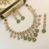 Gold-plated Indian Kundan jewelry set featuring a statement necklace, matching drop earrings, and a pair of teardrop-shaped earrings, decorated with light green stones and clear crystals, displayed on a cream-colored patterned book.