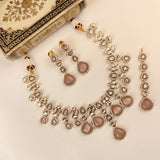 A traditional Indian jewelry set featuring a necklace, earrings, and a matching bracelet, styled on a light cream background with an ornate decorative box in the top left corner. The jewelry is made of silver-toned metal with clear stones and features dusty pink teardrop-shaped stones.