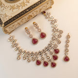 Gold-plated Indian jewelry set featuring a necklace, matching earrings, and a dangling piece, decorated with clear crystals and red heart-shaped stones, displayed next to an ornate cream-colored decorative box on a light beige background.
