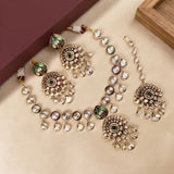 Gold-plated Indian Kundan jewelry set featuring a necklace, earrings, and a bracelet, embellished with clear crystals, green and purple enamel, and white stones, arranged on a beige surface.