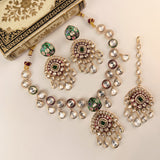 Gold-plated Indian jewelry set featuring a necklace, earrings, and a dangling maang tikka, embellished with clear crystals, green enamel, and red stone accents, displayed on a cream background next to an ornate decorative book.