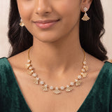 A close-up of a woman wearing a gold necklace and matching drop earrings with clear stones. The necklace features alternating gold fan-shaped and floral motifs, and the matching earrings are worn on her ears.
