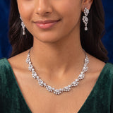 A close-up of a woman wearing a sparkling floral diamond necklace and matching drop earrings, styled with a dark green velvet top against a dark blue background.