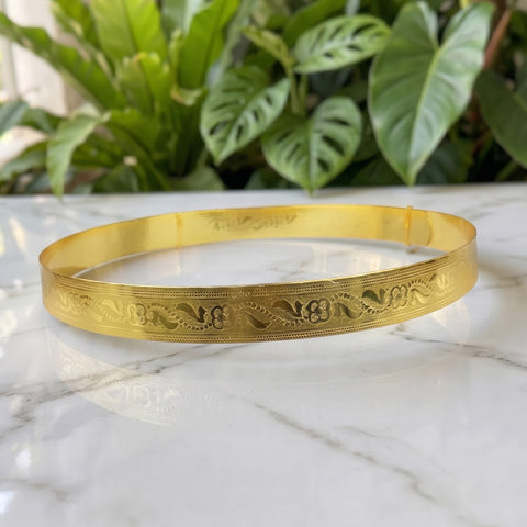 A gold-colored metal bangle bracelet with intricate floral engravings, resting on a white marble surface with green plants in the background.