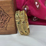 Decorative gold bangles with gemstones on a white surface with a wooden box and pink fabric in the background.