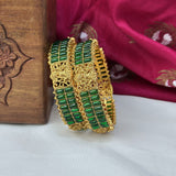Gold bracelet with green stones on a white surface with a wooden box and pink fabric in the background