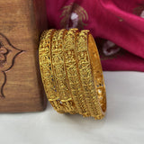 Gold bangles with intricate designs on a white surface with a wooden box and pink fabric in the background.