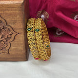 Gold bangles with green stones on a white surface with a wooden box and red fabric in the background