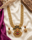 An ornate gold-toned traditional necklace with matching earrings, decorated with intricate multicolor enamel details in green, pink, and red, displayed on a cream satin fabric with purple silk and dried floral props.