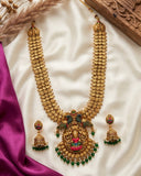 An ornate gold-toned traditional necklace with matching drop earrings, decorated with green and pink enamel details and set on a white satin fabric with purple silk and dried floral props.