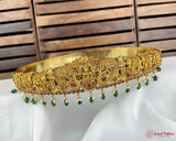 Gold necklace with green and red stones on a white surface with wooden background